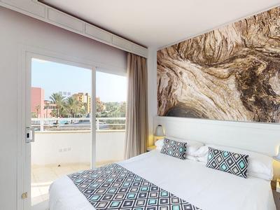 Jasny pokój hotelowy z łóżkiem podwójnym, abstrakcyjną sztuką na ścianie i balkonem z widokiem na zewnątrz.