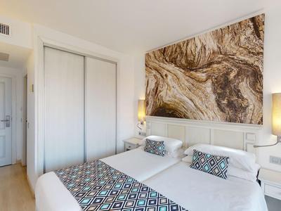 Jasny pokój hotelowy z podwójnym łóżkiem, dekoracyjnymi poduszkami i dużą abstrakcyjną sztuką na ścianie.