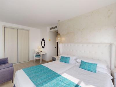 Jasny pokój hotelowy z dużym łóżkiem, turkusowymi akcentami, fotelami i nowoczesnym wystrojem.