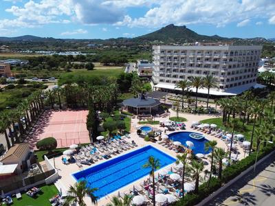 Hotel with large pool, tennis court, and mountains in the background under blue sky.