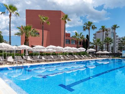 Grande piscine d'hôtel bleue avec chaises longues et parasols blancs, entourée de palmiers et d'un bâtiment rouge.