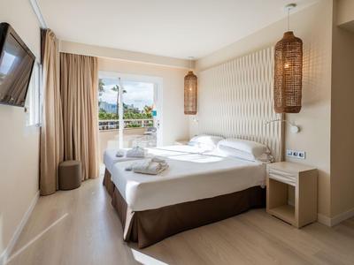 Bright hotel room with double bed, TV, slatted wall, and view of balcony and pool.