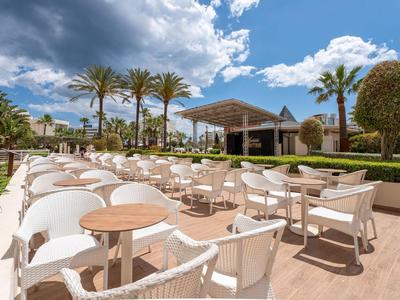 Café en plein air avec chaises blanches et tables devant des palmiers et un ciel bleu.