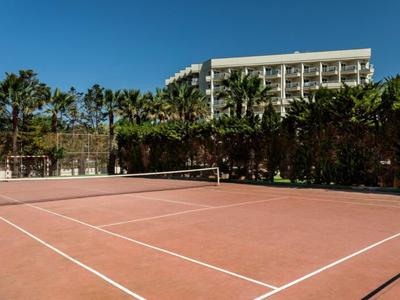 Court de tennis rouge avec des palmiers et un grand bâtiment hôtelier en arrière-plan sous un ciel clair.