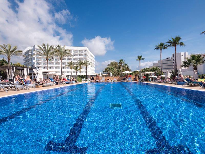 Duży basen na świeżym powietrzu w słonecznym kurorcie z palmami i leżakami.