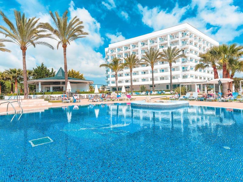 Basen w ośrodku z leżakami, parasolami, palmami i białym budynkiem hotelu w tle.