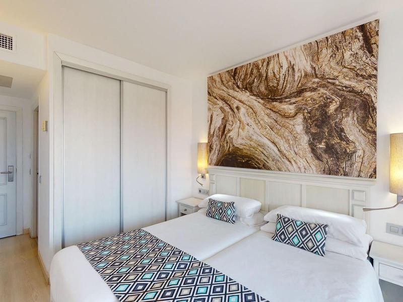 Jasny pokój hotelowy z podwójnym łóżkiem, dekoracyjnymi poduszkami i dużą abstrakcyjną sztuką na ścianie.