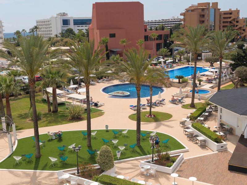 Strefa basenu hotelowego z palmami, leżakami, parasolami i pobliskimi budynkami.