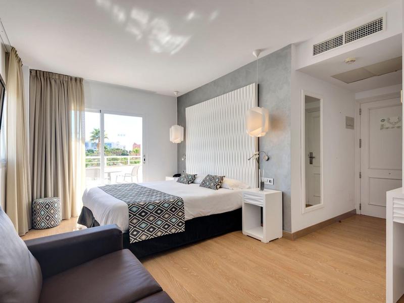 Jasny pokój hotelowy z podwójnym łóżkiem, sofą, drewnianą podłogą i dużym oknem z zasłonami.