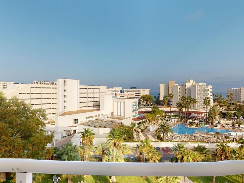 Duży hotel z basenami, palmami i widokiem na ocean pod jasnym niebem.