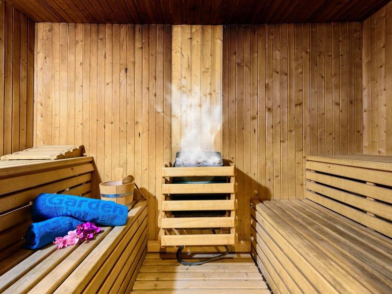 Sauna z drewnianymi ścianami, parujący piec, drewniane ławki, niebieskie ręczniki i różowe kwiaty.