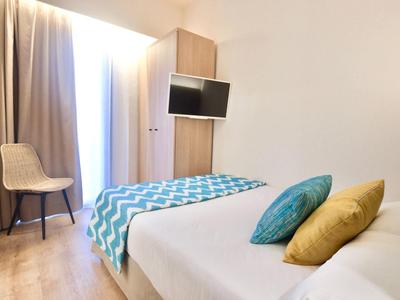 Helles Schlafzimmer mit schlichtem Bett, bunten Kissen, Fernseher an Holzschrank und Stuhl an Fenster.