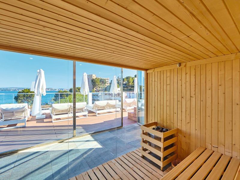 Sauna room with wooden paneling and a view of a terrace with loungers and the sea.