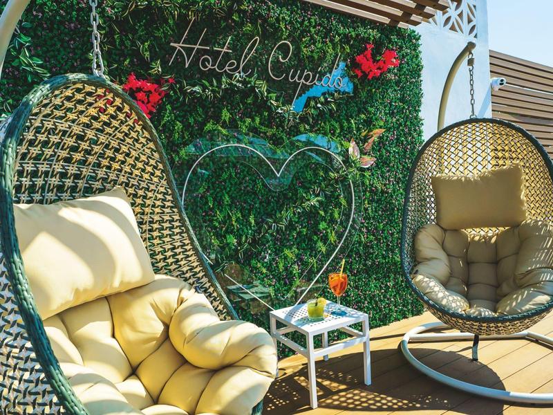 Two wicker chairs with cushions on a terrace in front of a decorative green wall with hotel name.