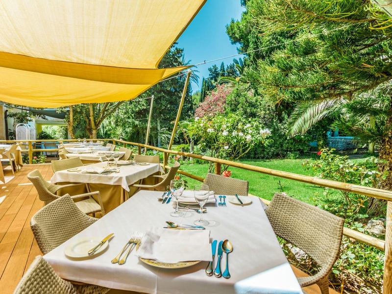 A set outdoor table with multiple chairs under yellow sunshades next to a garden.