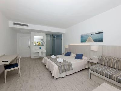 Habitación de hotel luminosa con cama doble, sofá cama, escritorio y baño moderno al fondo.