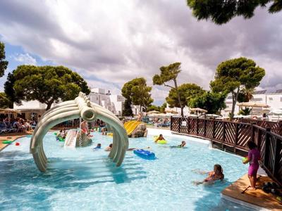 Área de piscina de hotel con tobogán y huéspedes, rodeada de edificios blancos y árboles bajo cielo nublado.