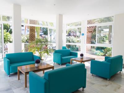 Área de salón de hotel luminosa con grandes ventanales, sofás turquesas y mesas de madera.