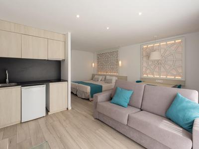 Habitación de hotel moderna con kitchenette, sofá y cama en tonos neutros con acentos azules.