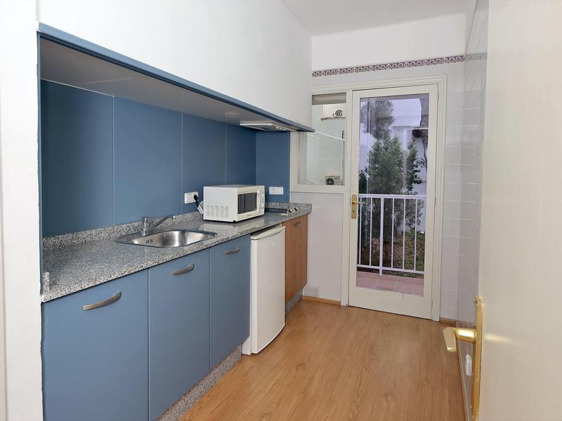 Pequeña cocina de hotel con armarios azules, microondas, fregadero y balcón al fondo.