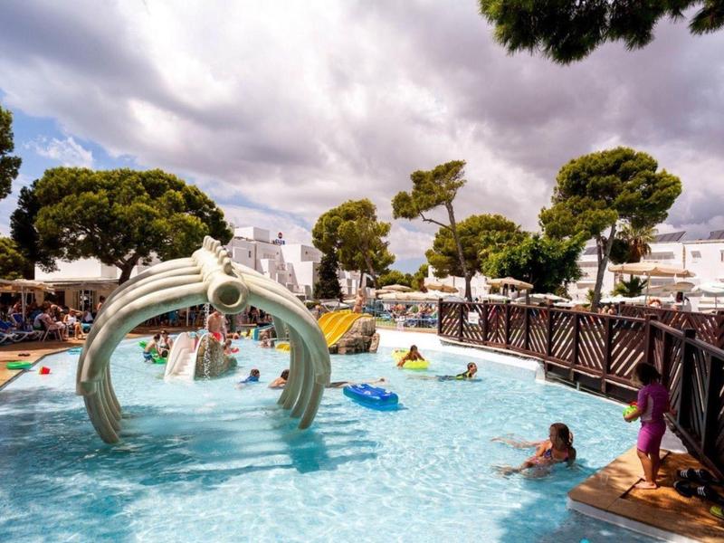 Área de piscina de hotel con tobogán y huéspedes, rodeada de edificios blancos y árboles bajo cielo nublado.