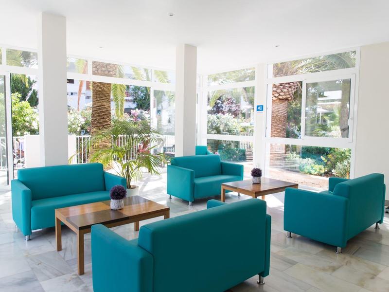 Área de salón de hotel luminosa con grandes ventanales, sofás turquesas y mesas de madera.