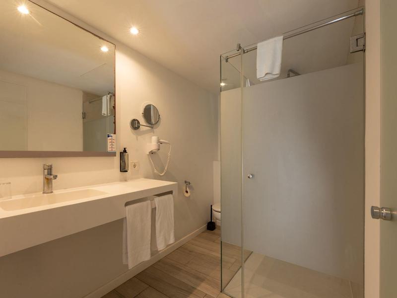 Baño moderno con gran espejo, lavabo, toallas y ducha de vidrio con luz cálida.