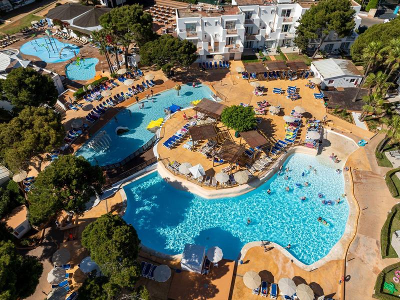 Vista de una gran zona de piscinas de hotel con tumbonas y personas en el agua bajo un sol brillante.