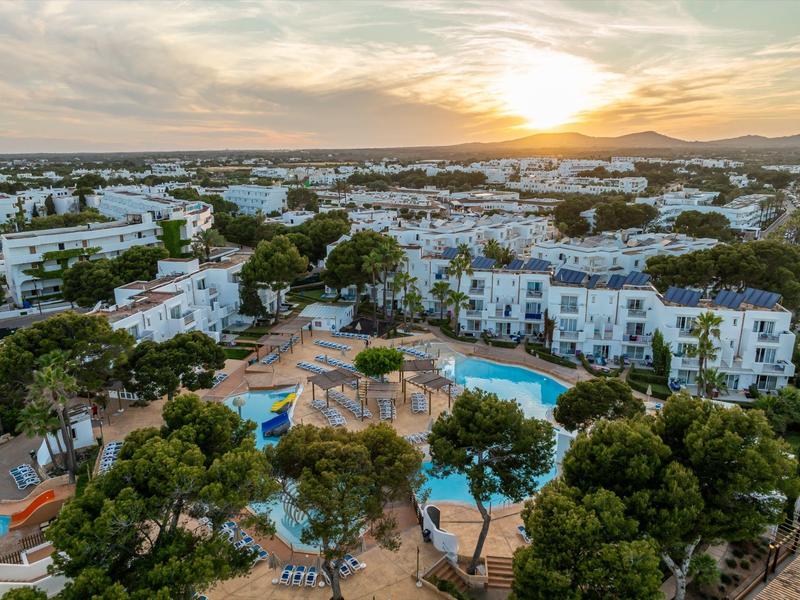 Vista aérea de un hotel con piscina rodeado de árboles y edificios blancos al atardecer.