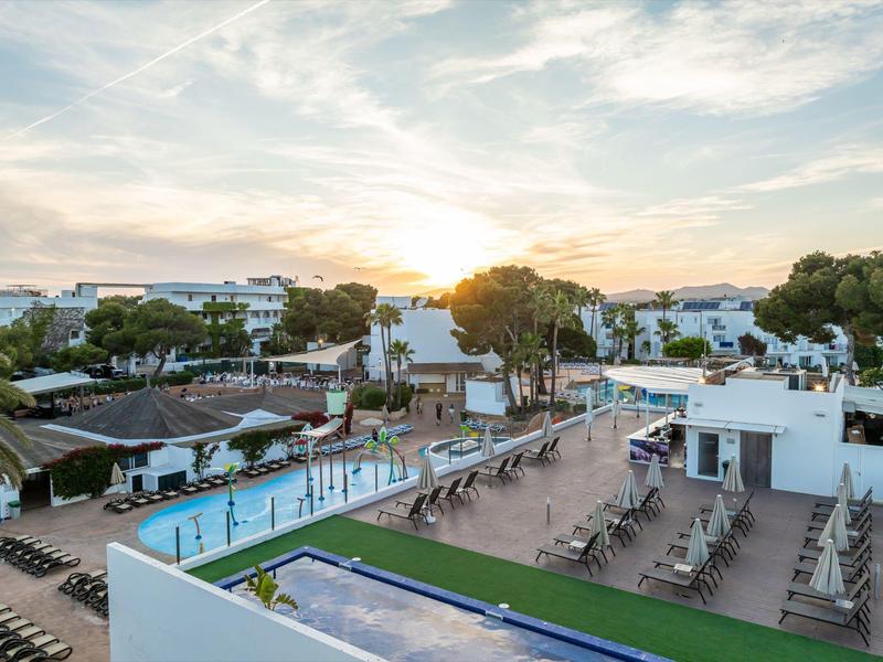 Complejo hotelero con piscina, tumbonas y edificios blancos bajo un cielo nublado al atardecer.