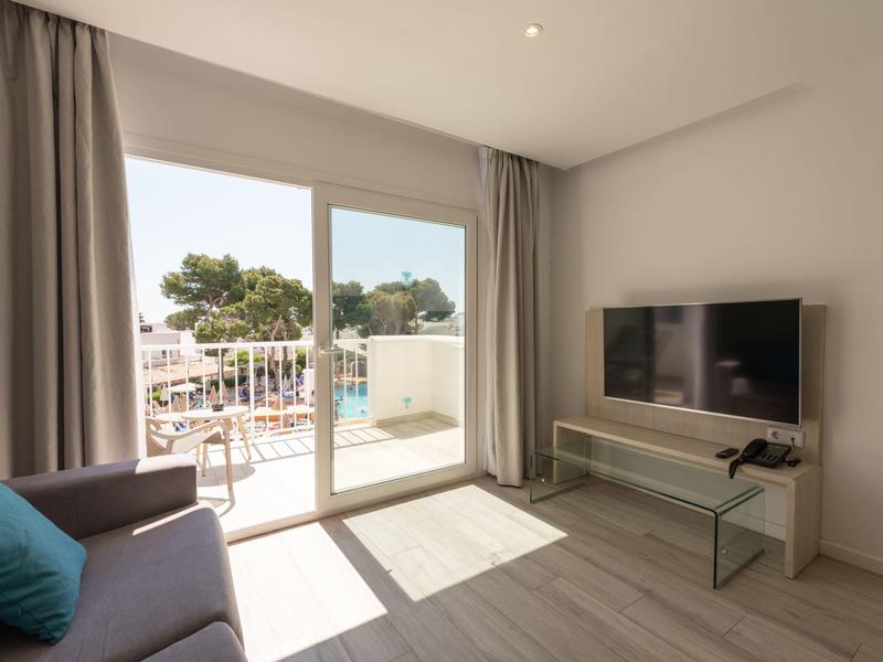 Habitación de hotel luminosa con sofá, TV y vista a terraza con piscina y árboles.