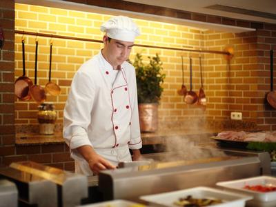Ein Koch in weißer Uniform arbeitet vor einem dampfenden Herd in einer Küchenumgebung mit Backsteinwand.