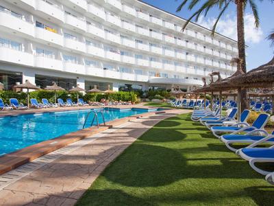 Großer Außenpool mit blauen Liegestühlen und Palmen vor einem weißen Hotelgebäude.