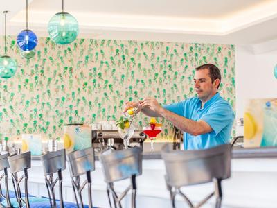 Ein Barkeeper mixt Getränke in einer modernen Bar mit grüner Tapete und blauen Hängelampen.