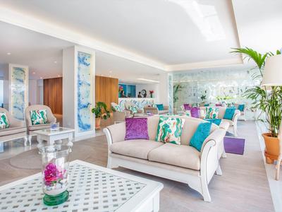 Helles Wohnzimmer mit weißen Möbeln, bunten Kissen, Glasvase und großen Zimmerpflanzen.