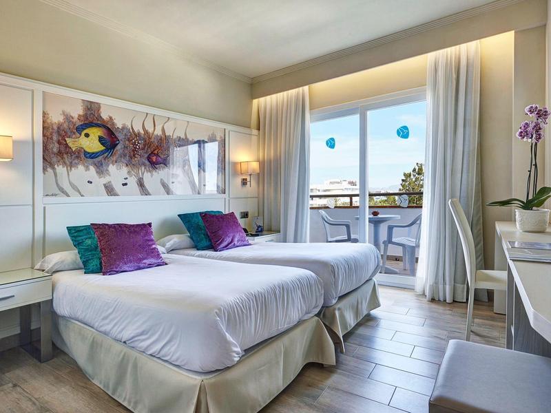 Helles Hotelzimmer mit zwei weißen Betten, bunten Kissen, Balkon und modernem Dekor.