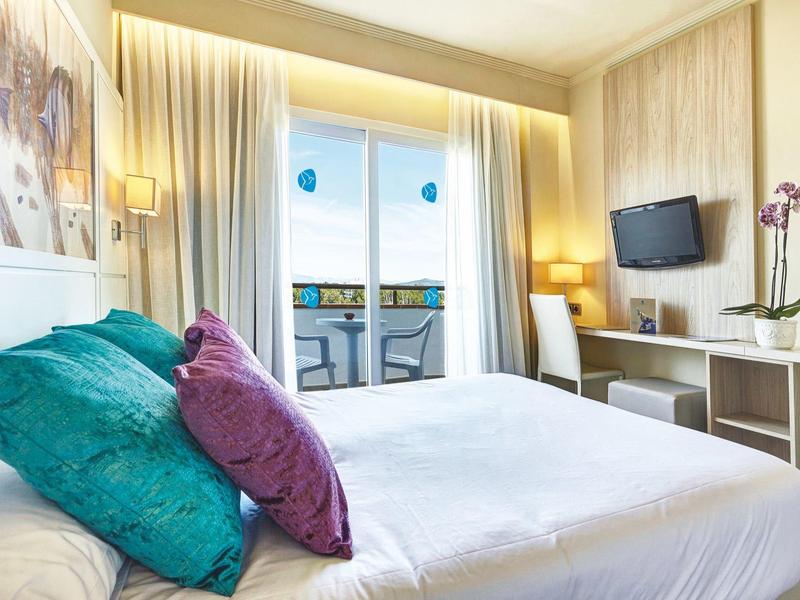 Modernes Schlafzimmer mit großem Bett, bunten Kissen, Balkon und weißem Schreibtisch mit Fernseher.