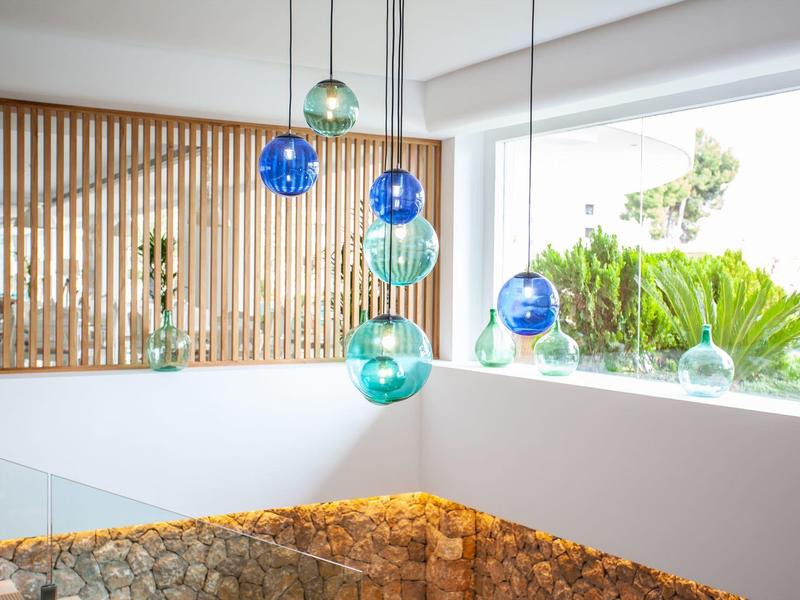 Mehrfarbige Glaslampen hängen über einem hellen, modernen Innenraum mit weißen Wänden und Holzlamellen.