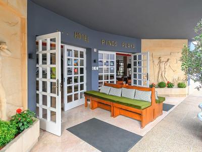 Área de entrada con puertas de vidrio abiertas, banco y plantas en un hotel.