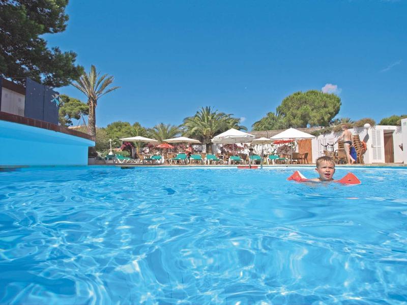 Woman swimming in clear pool water with sun umbrellas and trees in the background.