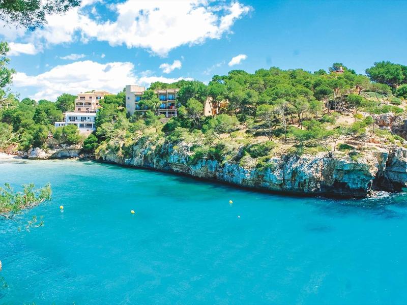 Clear blue water surrounded by rocky cliffs and green trees under a blue sky with clouds.