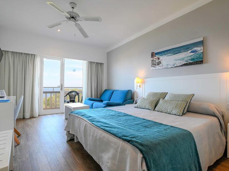 Bright hotel room with large bed, blue accents, and balcony with sea view.