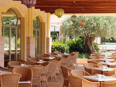 Überdachte Terrasse mit vielen geflochtenen Stühlen und Tischen, umgeben von Pflanzen und Bäumen.