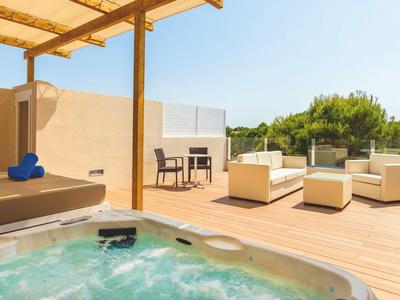 Terrasse mit Whirlpool, Lounge-Sofa, vier Stühlen und Grünflächen im Hintergrund bei Sonnenschein.