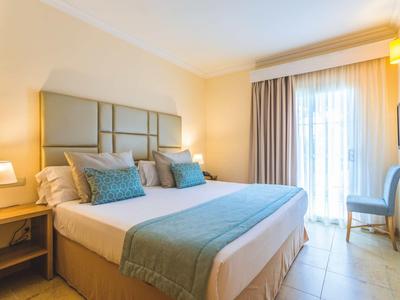 Helles Hotelzimmer mit Doppelbett, grauen Kissen, Nachttischen und lichtdurchflutetem Fenster mit Vorhängen.