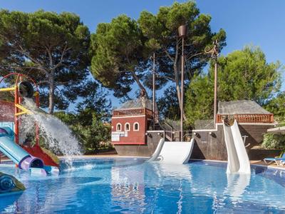Pool mit großen Wasserrutschen, Wasserspiel und Bäumen im Hintergrund bei klarem Himmel.