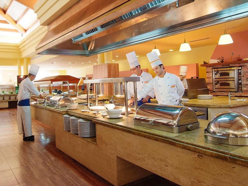 Buffetbereich mit zwei Köchen in weißer Kleidung und einer Person im Hintergrund, helle Holzdecke mit Tageslicht.