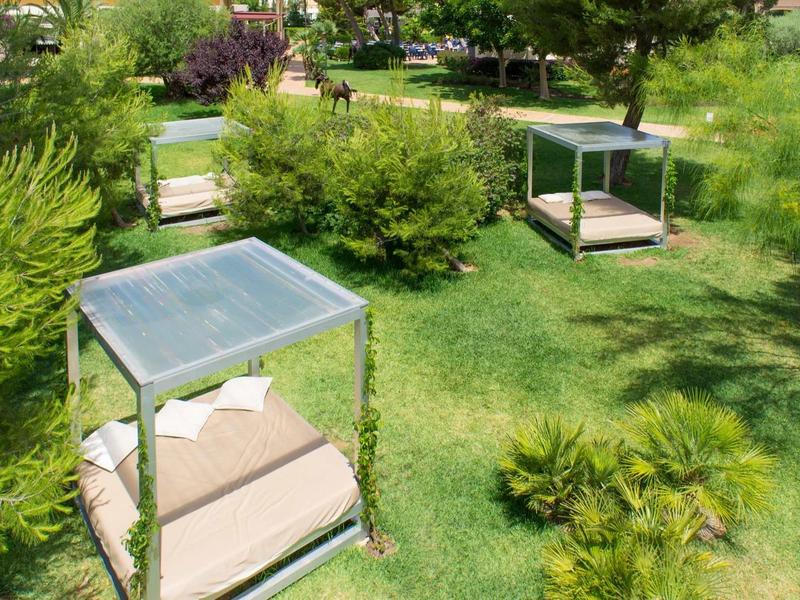 Grüne Parkanlage mit Gras, Büschen und vier modernen, quadratischen Sitzliegen mit Sonnenschutz.