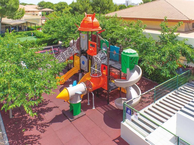 Bunter Spielplatz mit Rutschen und Klettergerüst, umgeben von Bäumen und Gebäuden.