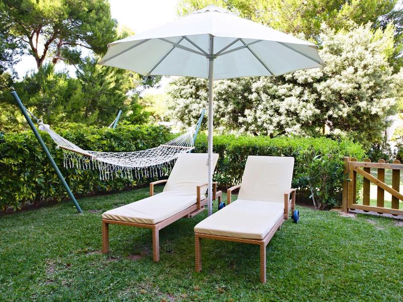 Grüne Gartenwiese mit zwei Holzloungebetten, weißem Sonnenschirm und Hängematte zwischen Bäumen.